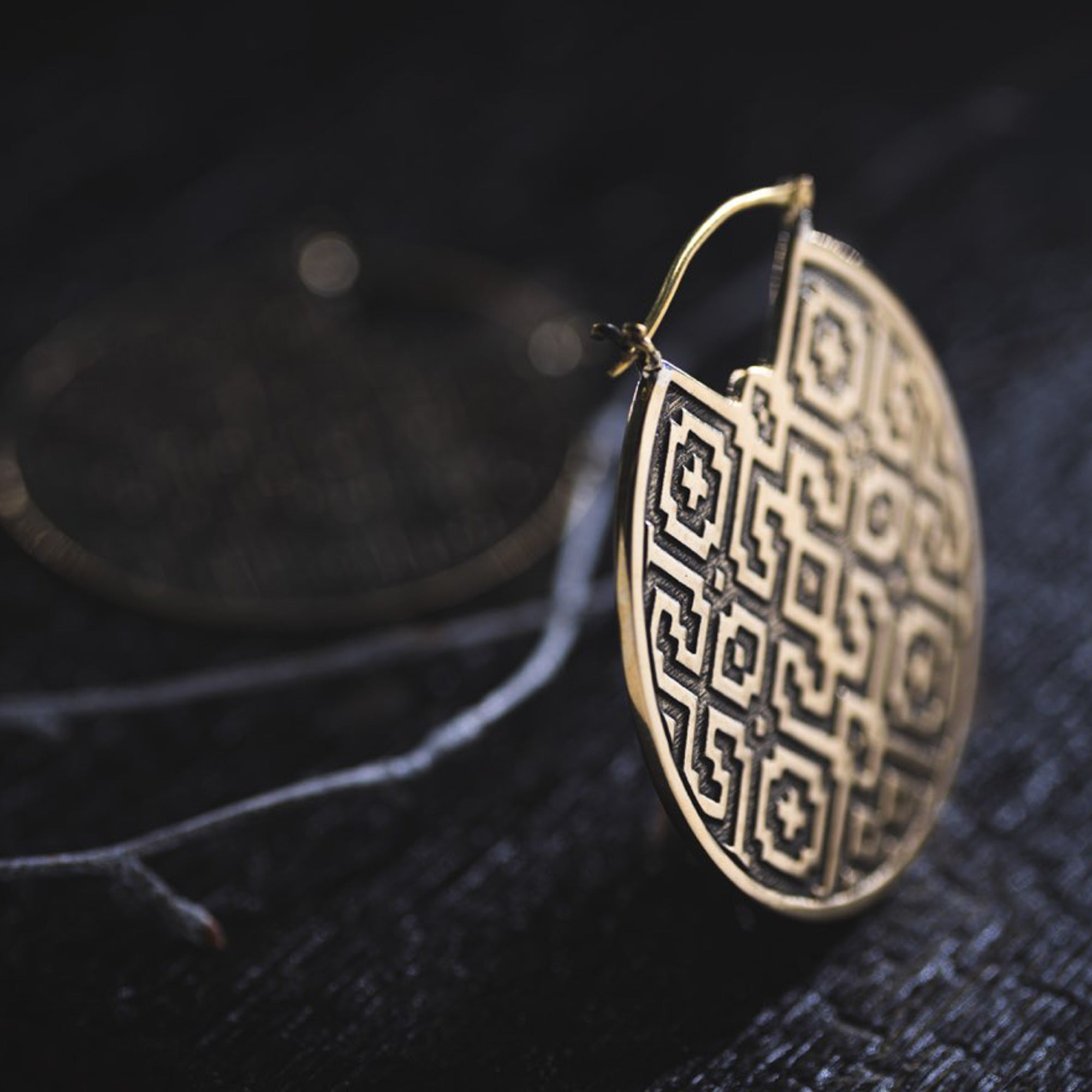 Close-up of a round, intricately patterned earring resting on a dark surface. The AREVALO Shipibo Oversized Disc Hoop Earring in Gold by Kaya Movement showcases sacred geometry Shipibo designs with repeating square and diamond shapes in a gold color. Another similar earring is blurred in the background, enhancing the tribal disc hoop earrings vibe.